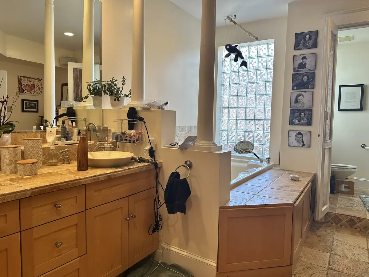 Before photo of a primary bathroom renovation in NW Washington, DC showing the original double-sink wood vanity, built-in bathtub, framed mirrors, and beige tile flooring.