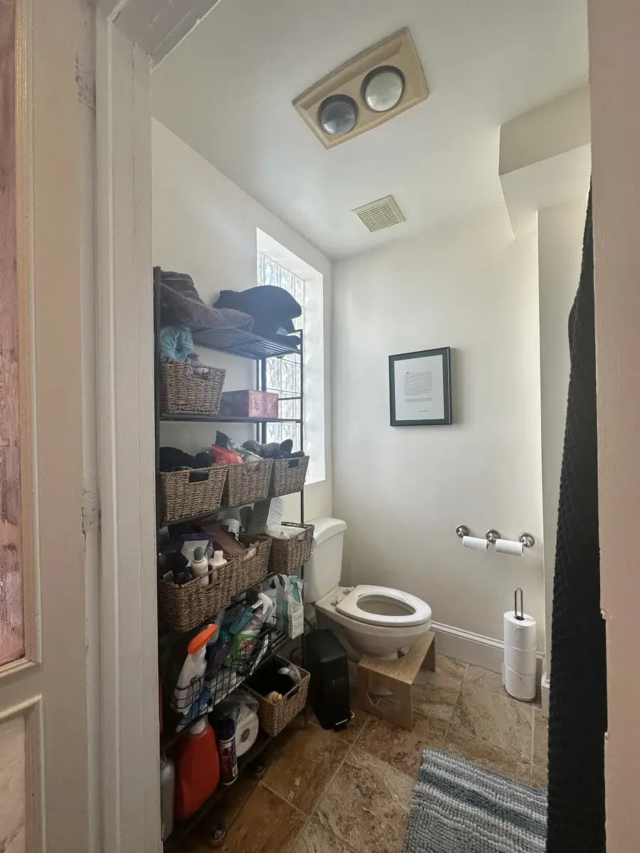 Before photo of a primary bathroom remodel in NW Washington, DC showing a narrow toilet area with open shelving, older tile flooring, and dated fixtures.