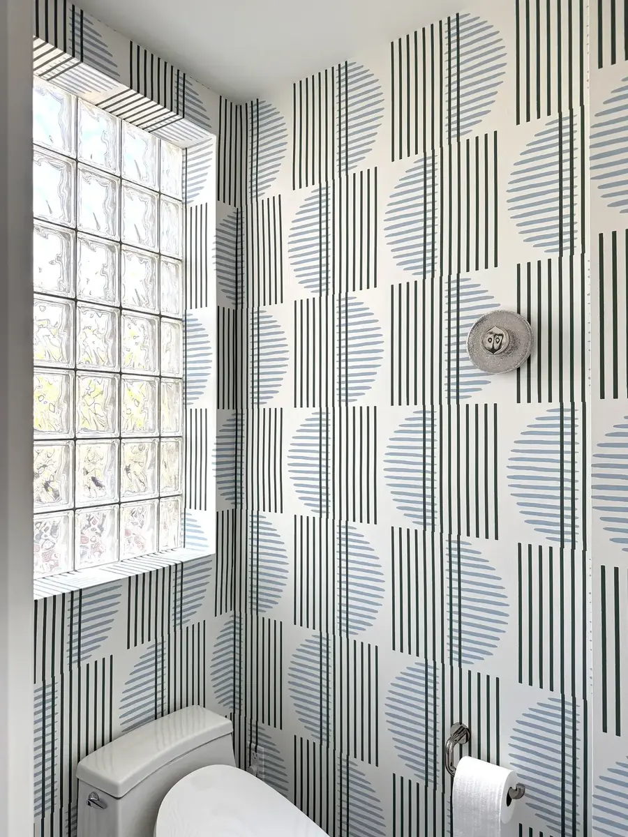 The toilet area in a NW Washington, DC primary bathroom remodel featuring graphic patterned wallpaper, white fixtures, and a glass block window.