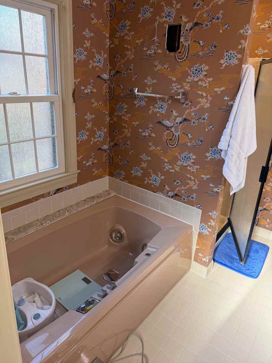Before photo of a primary bathroom remodel in Potomac, MD showing an outdated pink soaking tub, floral bird-patterned wallpaper, beige tile surround, and frosted window.
