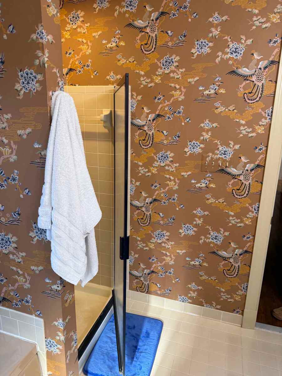 Primary bathroom before renovation in Potomac, MD featuring dated floral wallpaper, a framed glass shower enclosure, beige shower tiles, and white tile flooring.