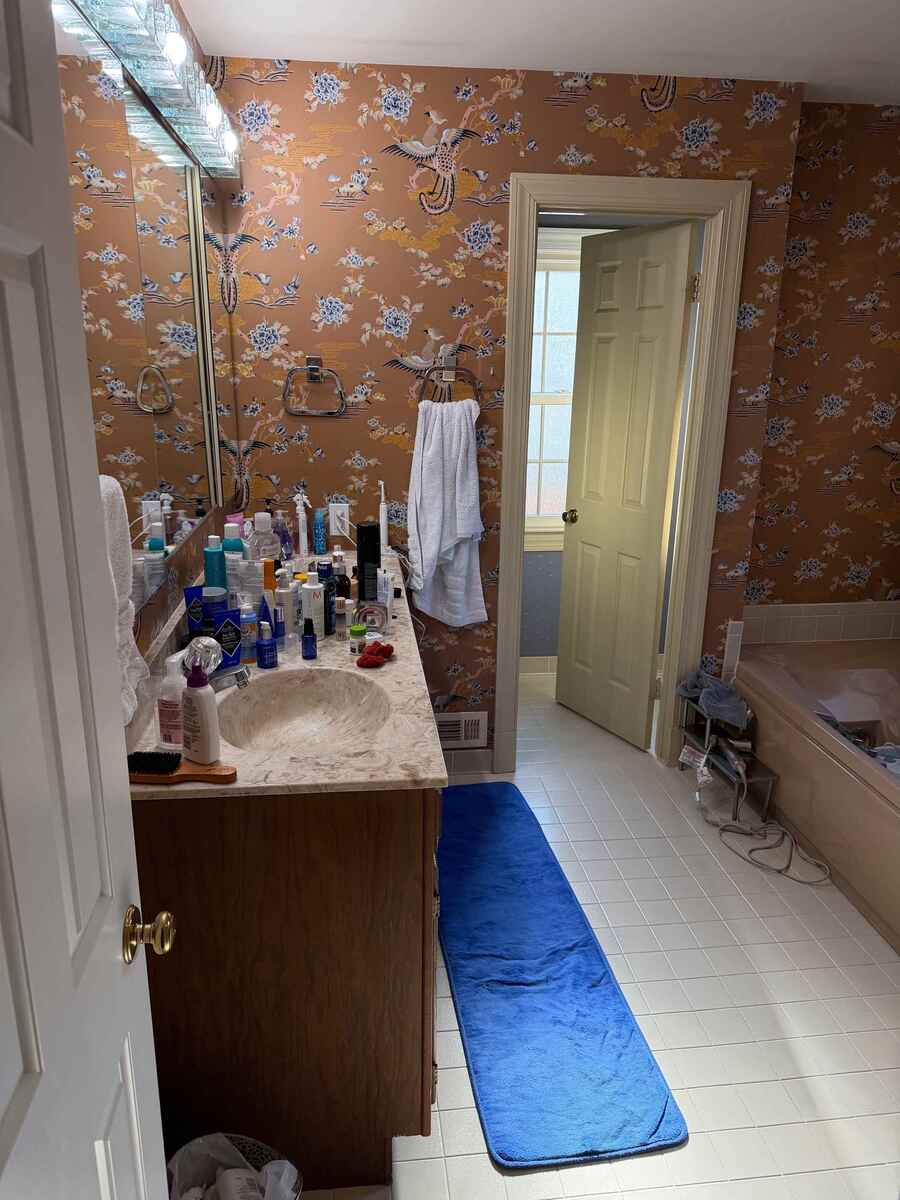 Before view of a primary bathroom renovation in Potomac, MD showcasing the original wood vanity, marble-look countertop, large wall mirror, pink bathtub, and patterned wallpaper.