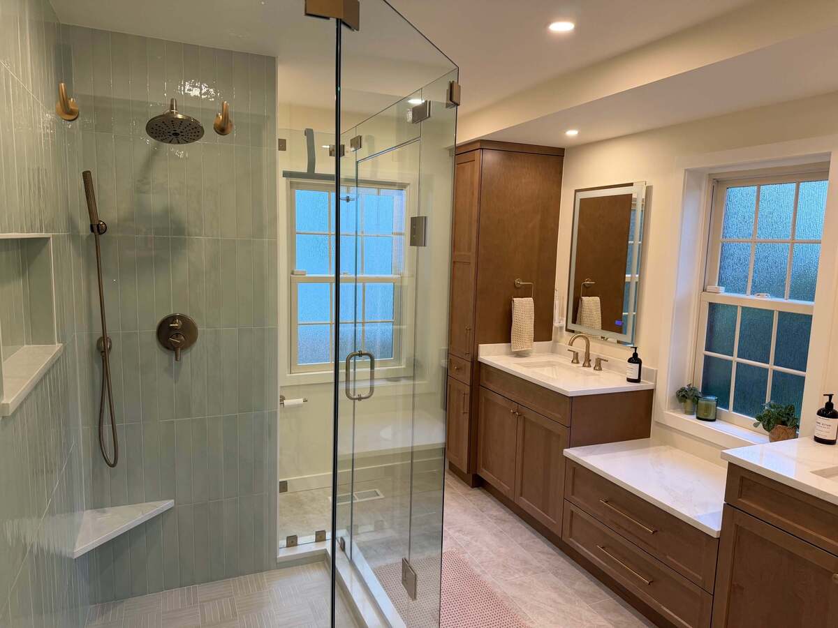 Remodeled primary bathroom in Potomac, MD with a spacious glass-enclosed shower, rainfall showerhead, warm wood vanity, quartz countertops, and built-in storage.