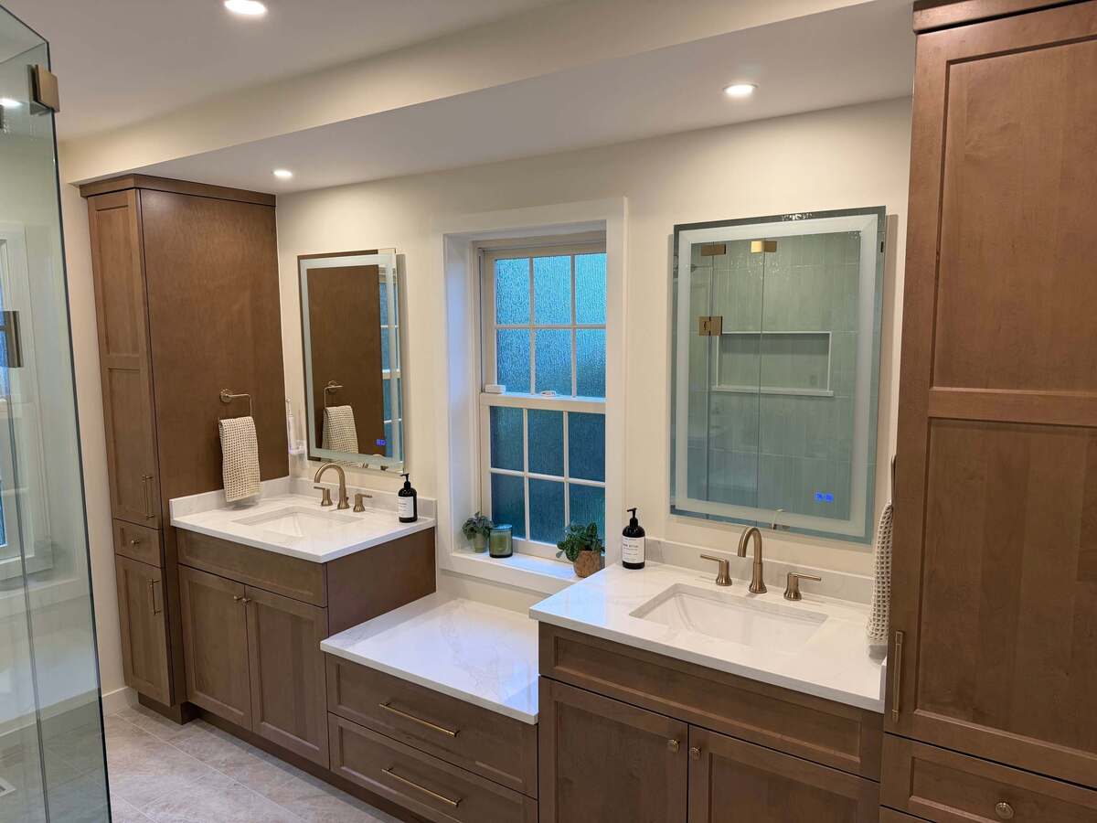 Custom double-sink bathroom vanity in Potomac, MD featuring rich wood cabinetry, white quartz countertops, brass faucets, and a built-in makeup counter beneath a window