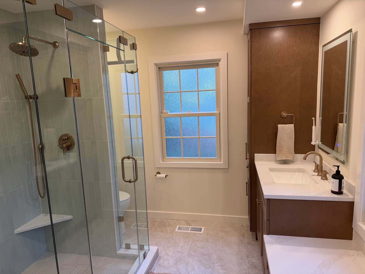 Primary bathroom remodel in Potomac, MD highlighting a frameless glass shower with sage green tile, a rainfall showerhead, custom wood cabinetry, and a frosted glass window.