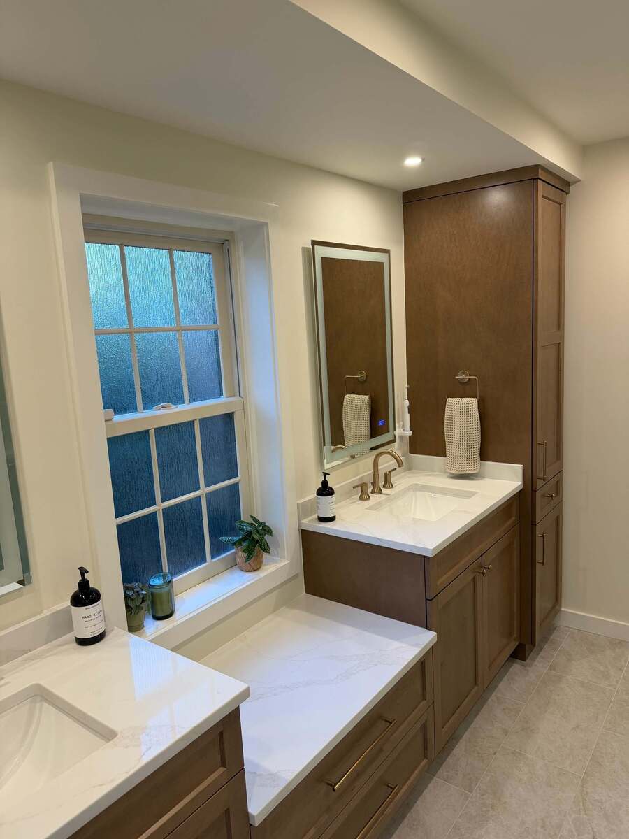 A bathroom vanity in a Potomac, MD bathroom remodel featuring custom wood storage cabinets, a white quartz countertop, undermounted sinks, and brushed brass fixtures.